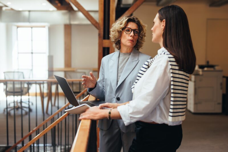 Two business women are having a discussion about patent application. They are standing on the first floor of an office, holding papers or a laptop in their hands.